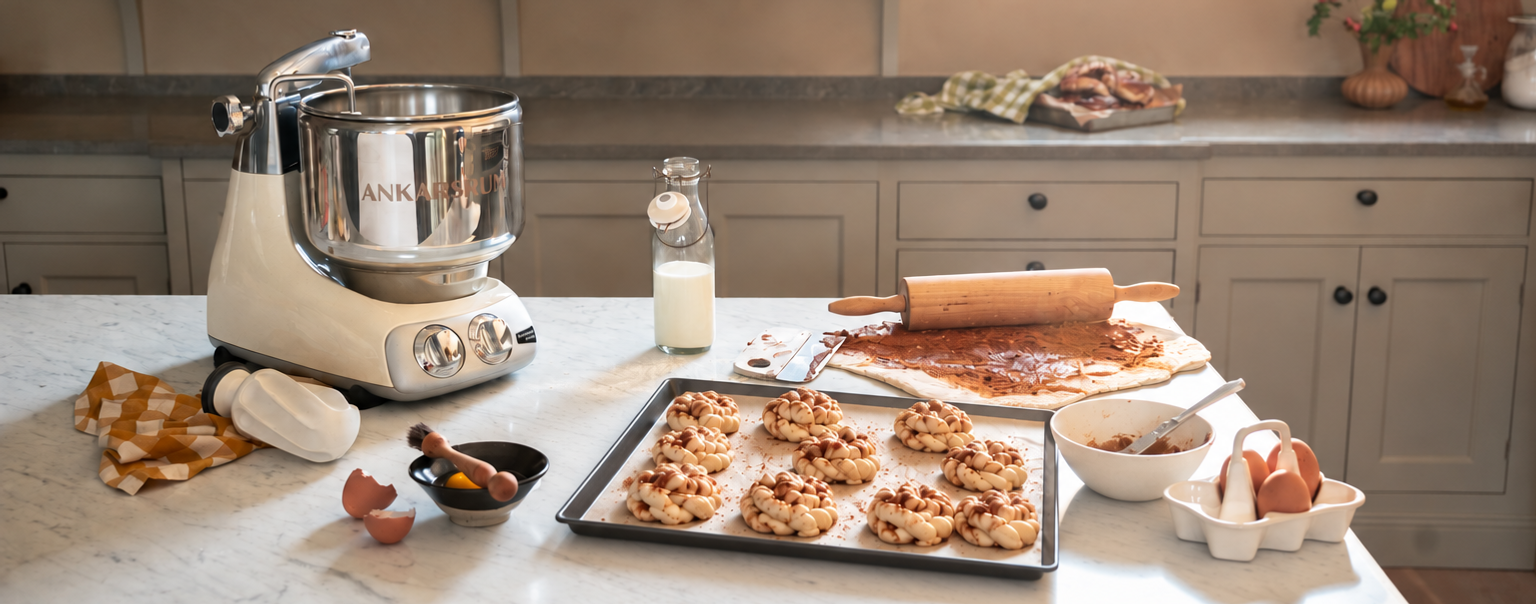 An Ankarsrum Assistent Original with a stainless steel bowl on a kitchen island, surrounded by freshly baked cinnamon buns on a baking tray and baking ingredients.