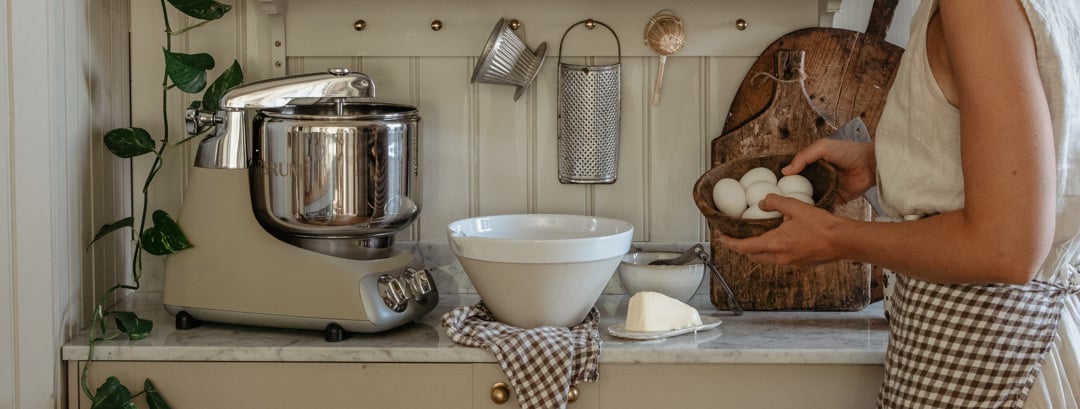 A woman prepares ingredients in a country-style kitchen beside an Ankarsrum Assistent Original in Harmony Beige.