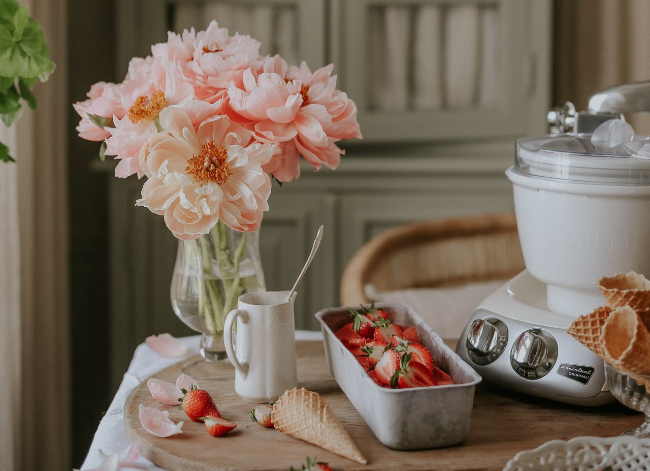 En skål med jordgubbar och en vas med blommor på ett bord. På bordet står en Ankarsrum Assistent Original i Cream, monterad med Glassmaskinen.