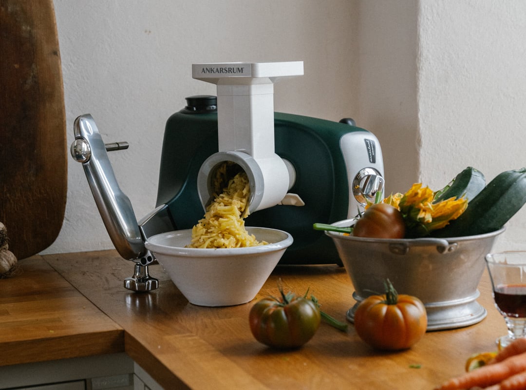 An Ankarsrum assistent original with a vegetable cutter attached shreds potatoes into a bowl.