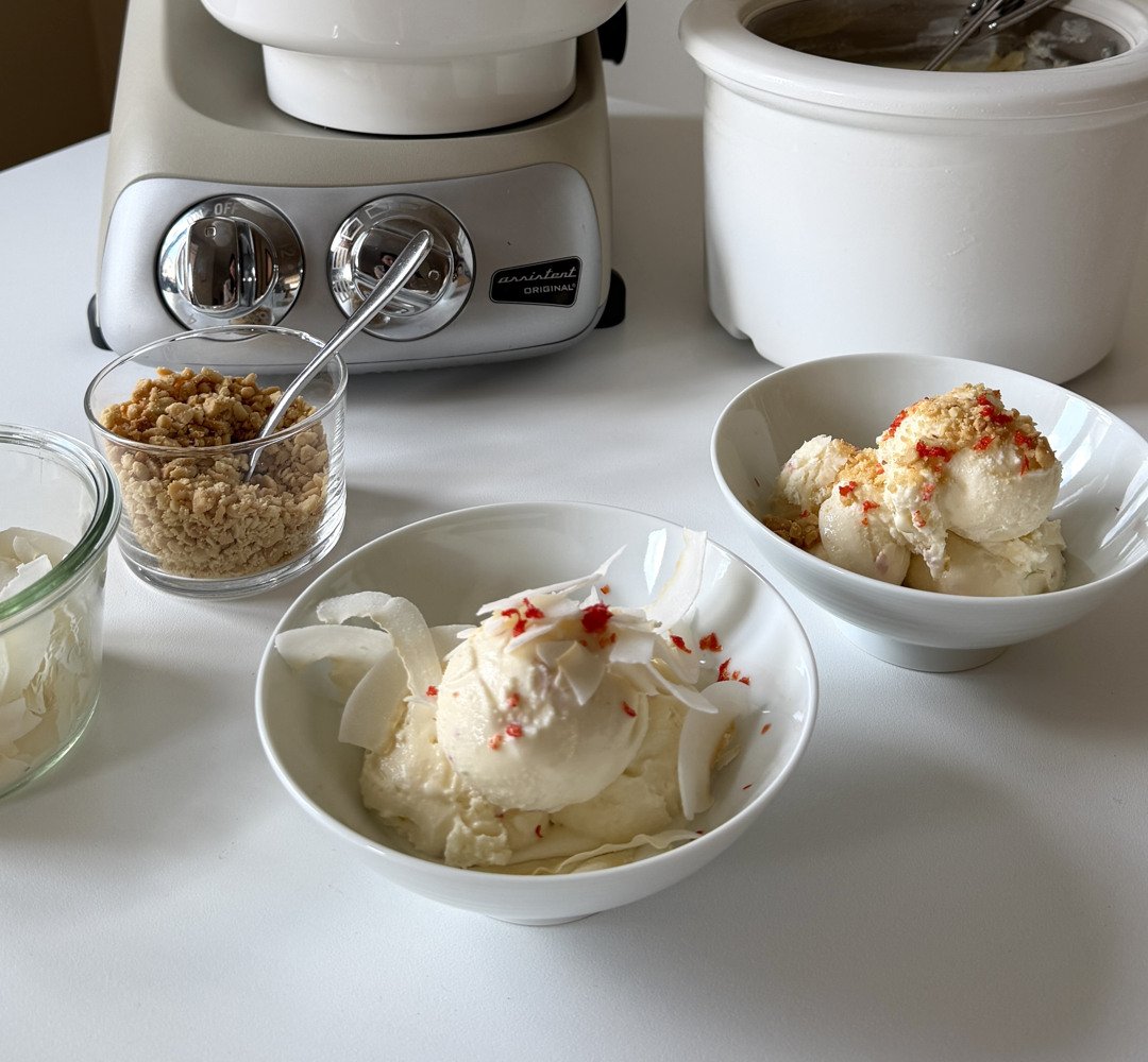 Ett vitt bord täckt med skålar av limeglass, garnerad med chili och citrongräs. I bakgrunden står en Ankarsrum Assistent Original i Harmony Beige utrustad med glassmaskins-tillbehöret.