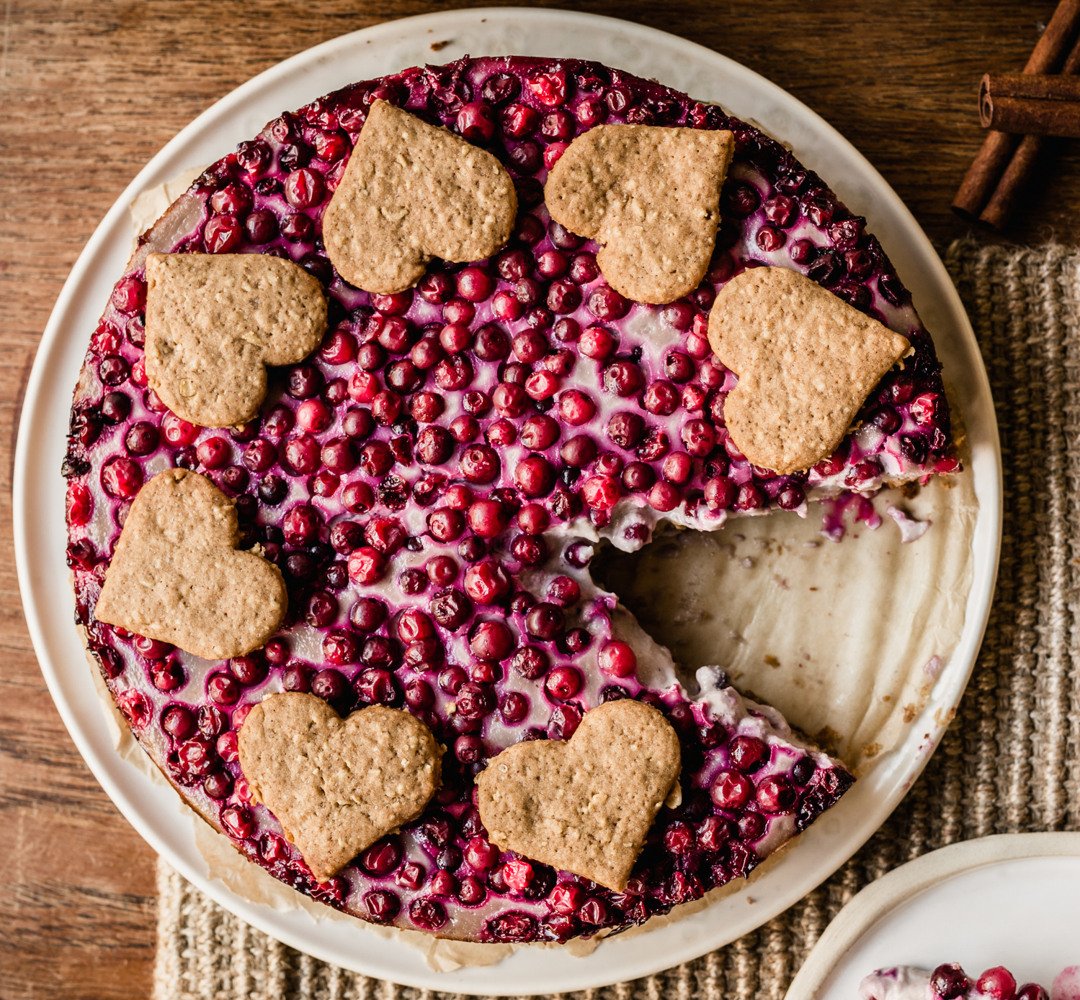 Cheesecake topped with lingonberries and gingerbread cookies on a wooden table.