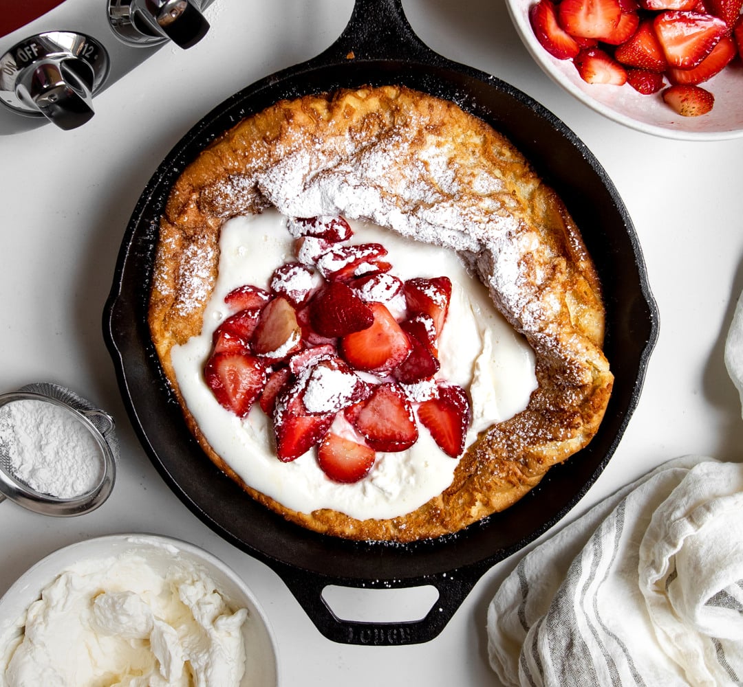 Dutch baby pancake with strawberries and cream in a cast iron skillet on a white table.