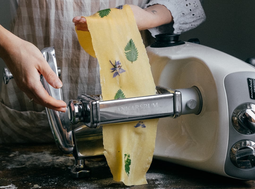A woman uses the Ankarsrum Pasta Roller attachment to roll fresh pasta dough decorated with edible flowers and herbs.