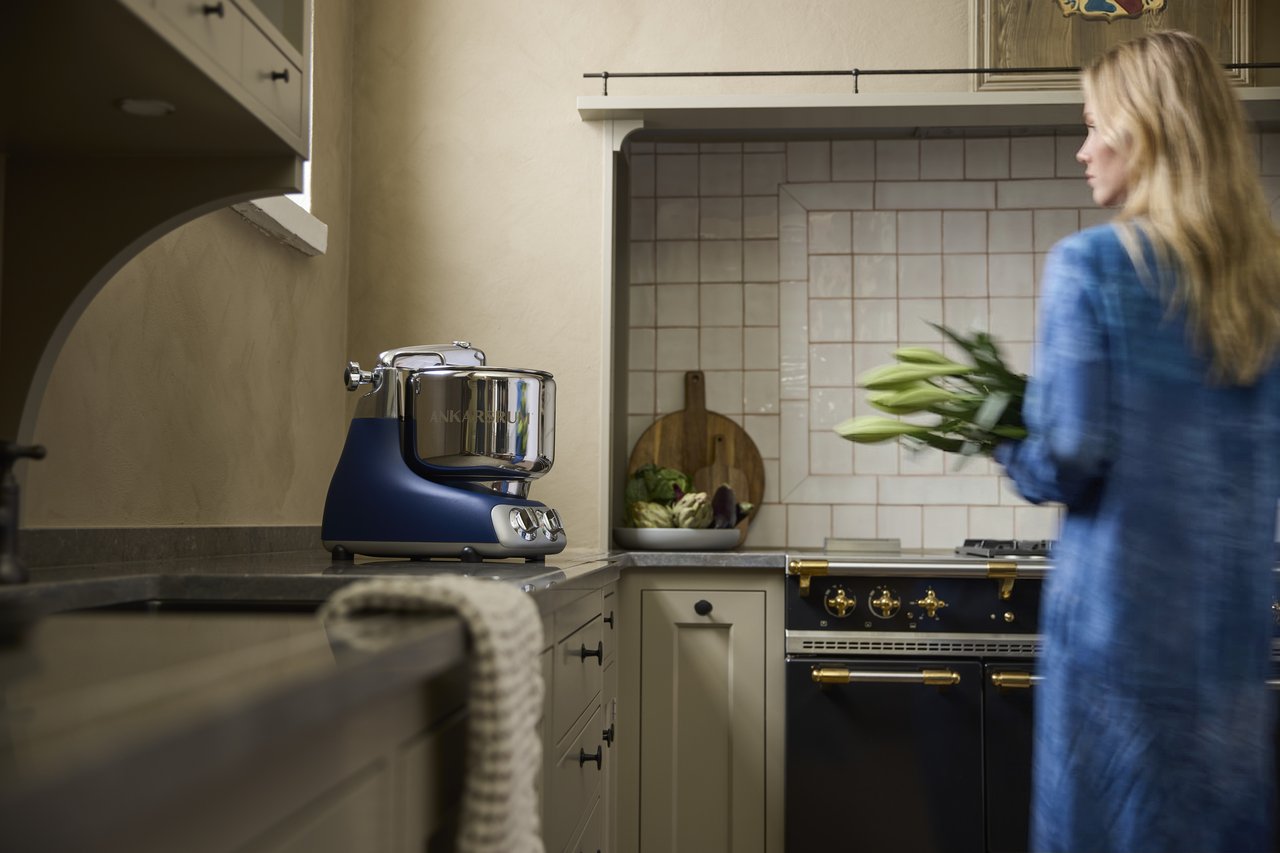 En kvinna i en blå klänning står i ett kök bredvid en Ankarsrum Assistent Original i Royal Blue.