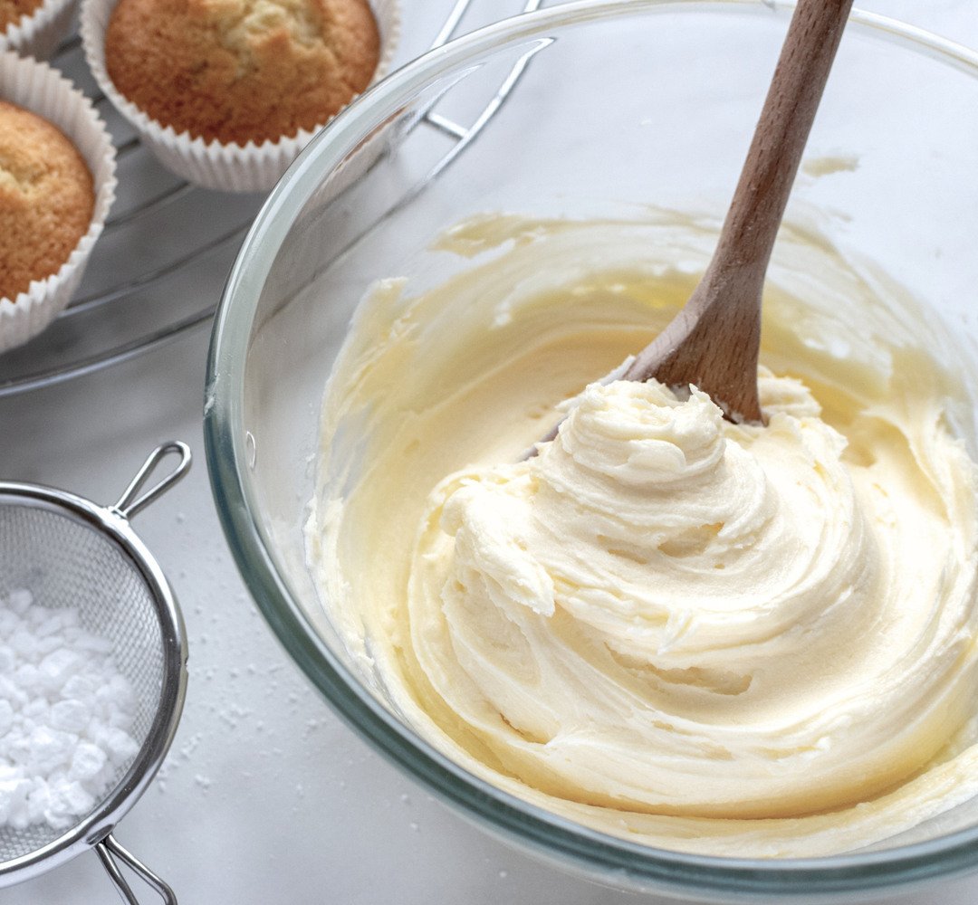 A glass bowl filled with buttercream next to muffins.
