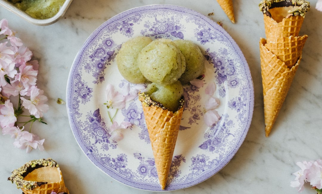 En person som håller en tallrik med en kiwi-sorbetkon. I bakgrunden syns rosa blommor, tillsammans med ett bord som har en skål med kvarvarande glass.
