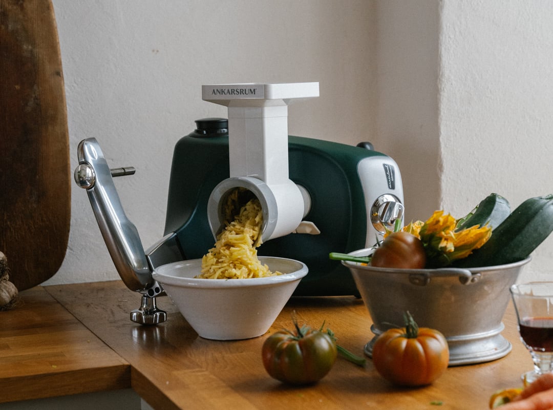 En Ankarsrum Assistent Original i Skogsgrön, utrustad med Grönsakskuttertillbehöret, står på en träbänk i köket omgiven av färska grönsaker och skivad zucchini.