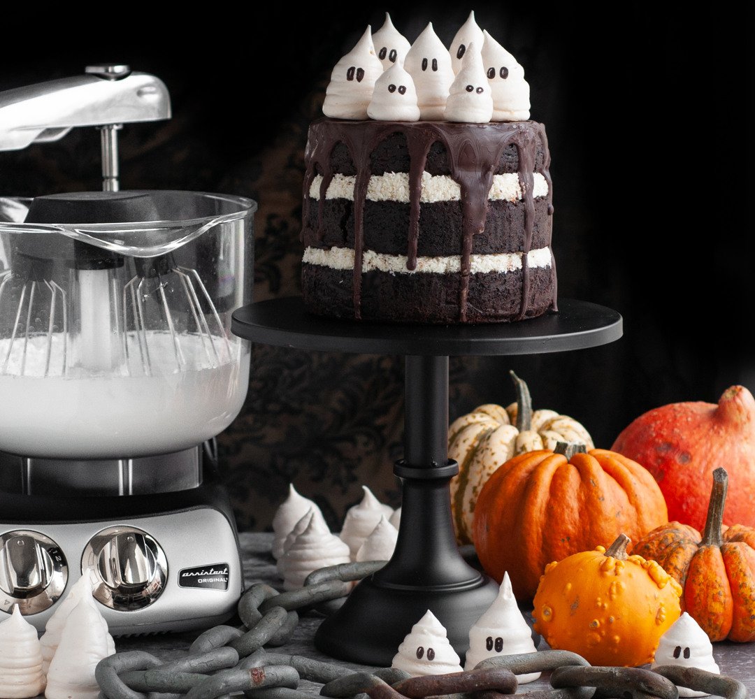A Halloween cake topped with meringue ghosts. An Ankarsrum Assistent next to the cake with the whisking bowl, surrounded by more meringue ghosts and pumpkins.
