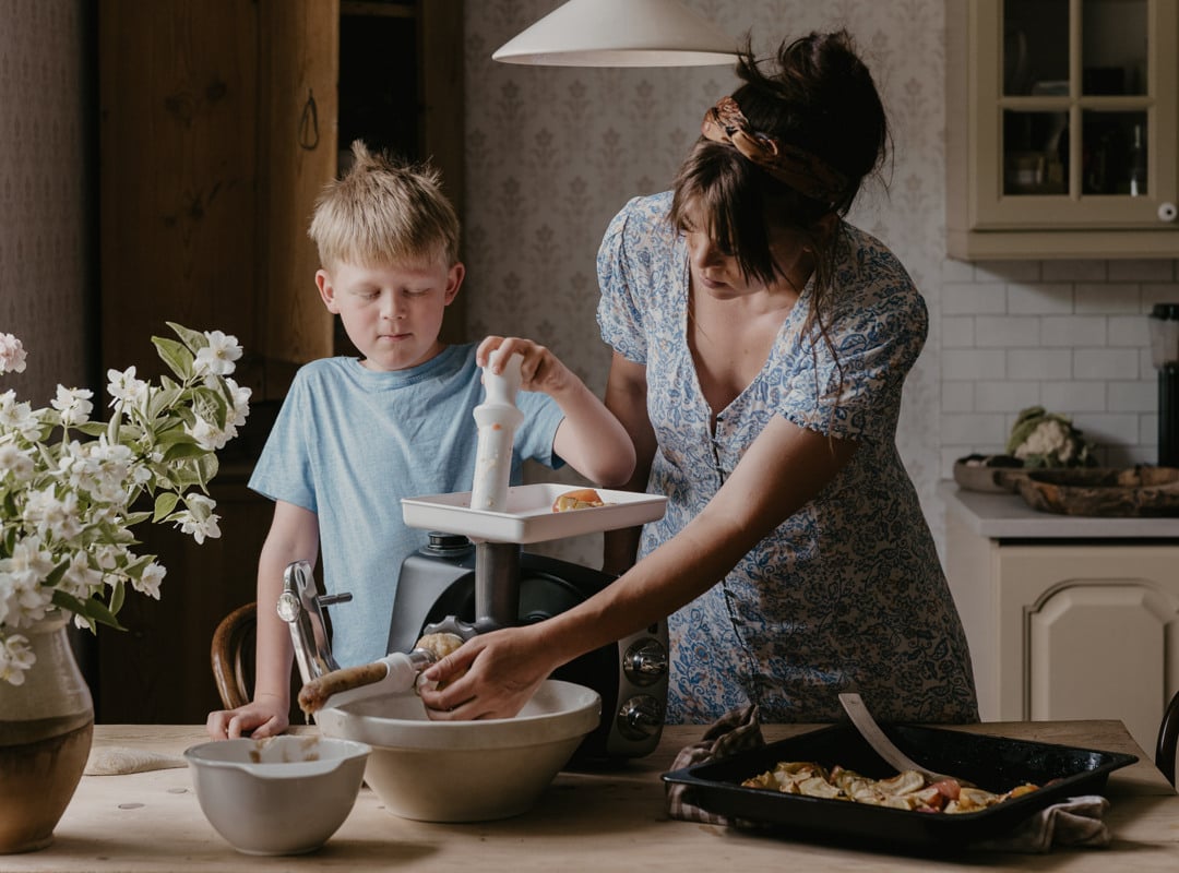 En kvinna och en pojke använder passertillsatsen på en Ankarsrum Assistent Original i färgen Forest Green för att göra färskt äppelmos i ett solbelyst kök.