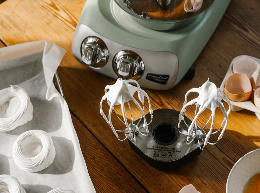 A pair of beaters covered in meringue besides an Ankarsrum stand mixer on a wooden table.