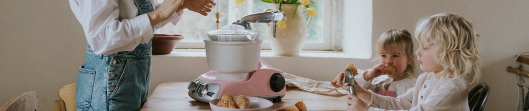 A woman and two children making ice cream together with a pink Ankarsrum Assistent Original at a wooden kitchen table.