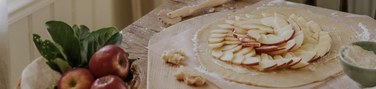 A rustic apple tart topped with thinly sliced apples on a table by a sunlit window, surrounded by fresh apples and baking ingredients.