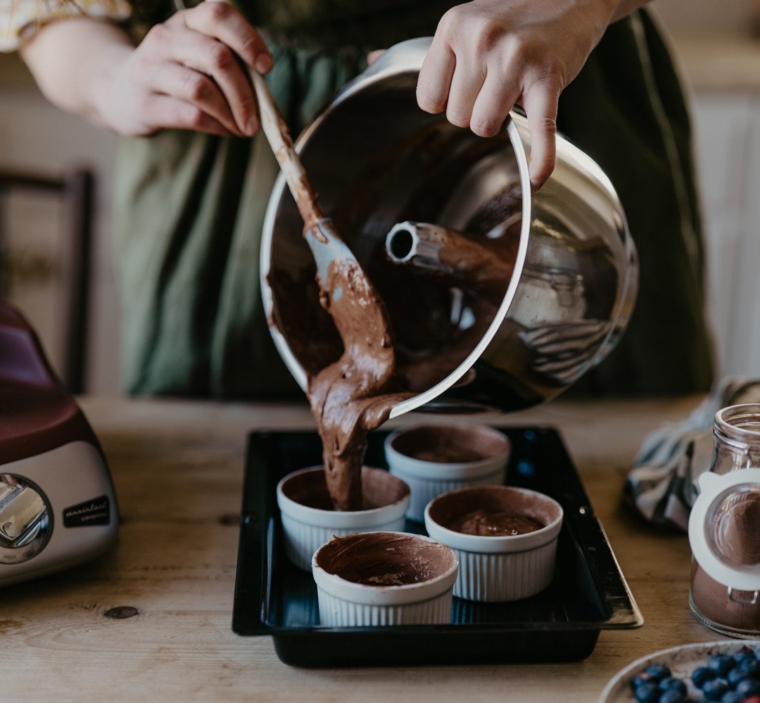 En kvinna iklädd ett grönt förkläde använder en slickepott för att hälla ut chokladfyllning ur en vispskål i rostfritt stål. Fyllningen hälls ner i vita bakformar som står på en plåt.
