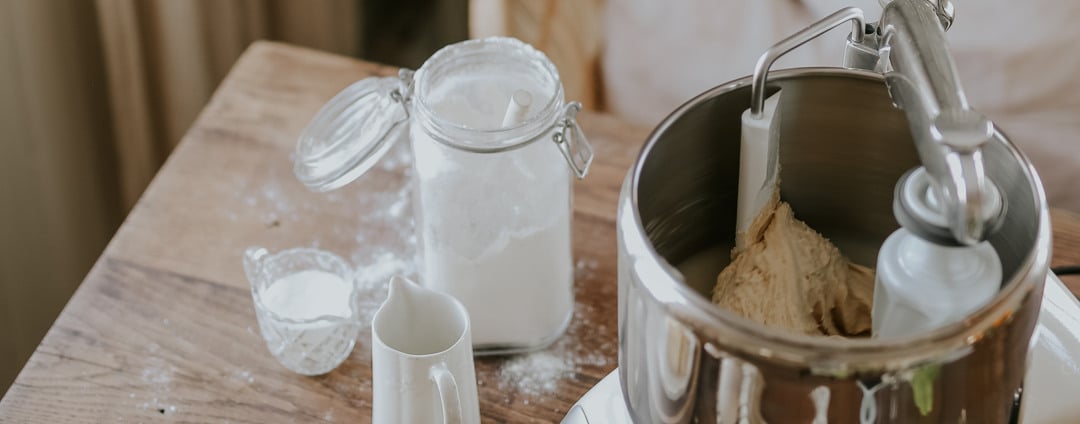 Close-up of bread dough being kneaded in an Ankarsrum Assistent Original, with flour and baking ingredients on a wooden table.