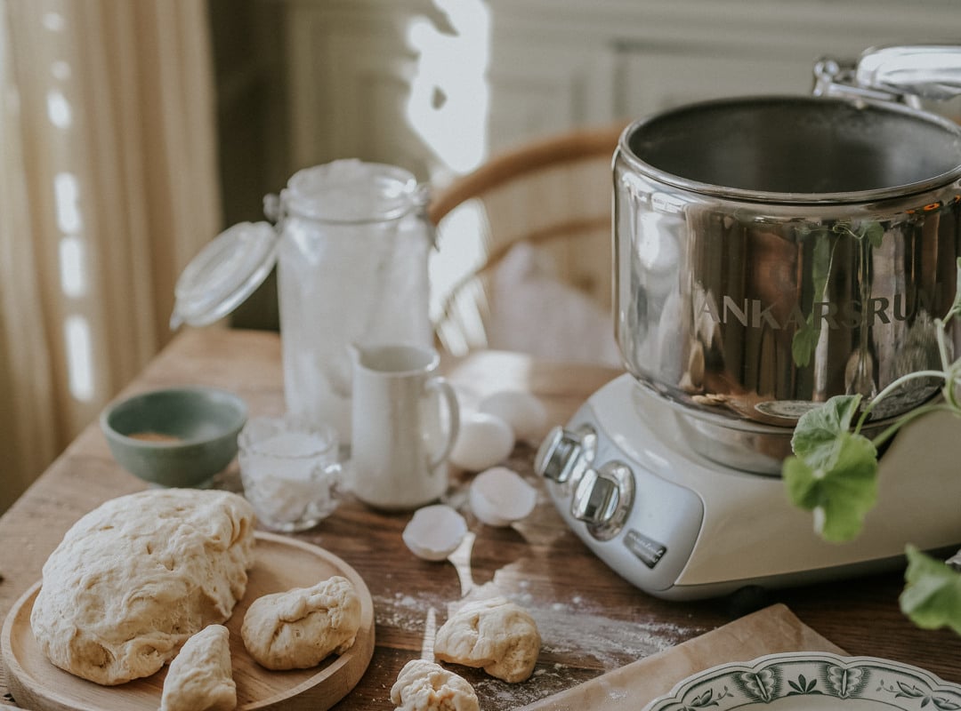 An Ankarsrum Assistent Original on a kitchen table surrounded by dough, freshly baked rolls and baking ingredients.