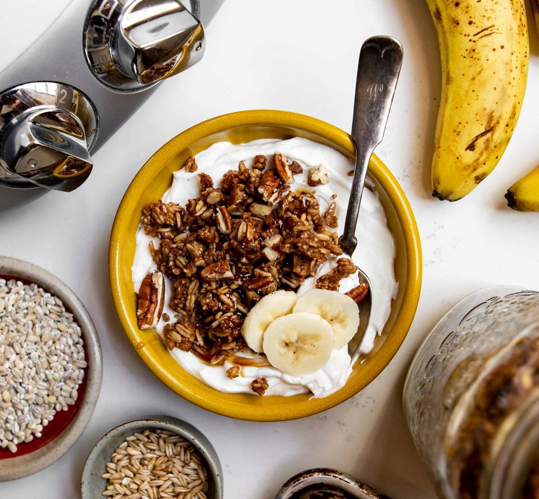 A bowl of yogurt, topped with homemade banana bread granola, on a table.