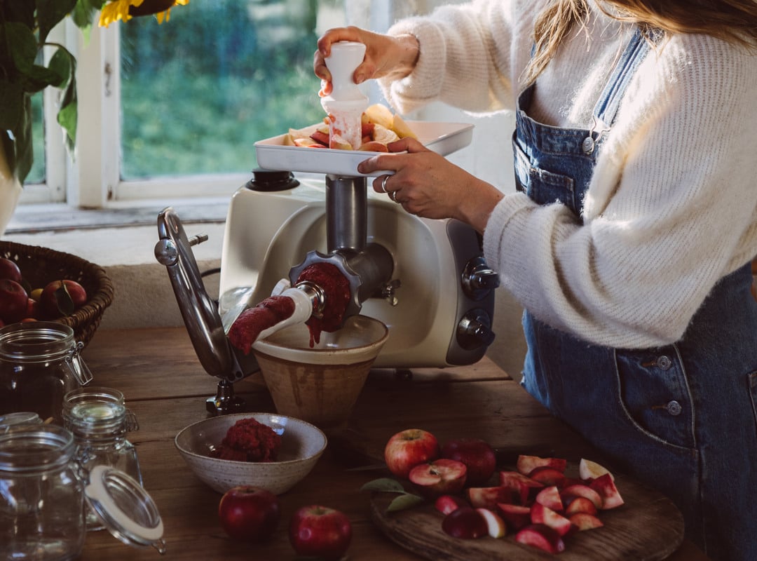 Nainen käyttää Strainer-liitintä Ankarsrum Assistent Originalissa Harmony Beige -värissä valmistellessaan tuoretta omenasosetta auringonvaloisassa keittiössä.