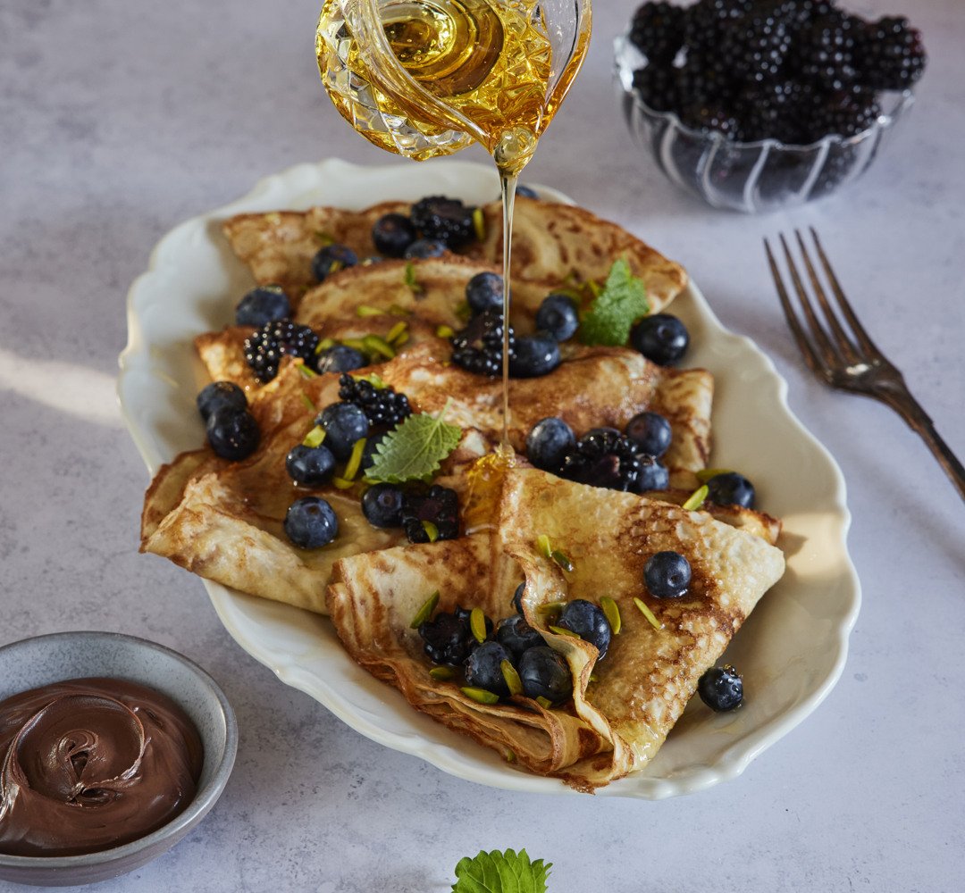 Crêpes mustikoiden, siirapin ja karhunvatukoiden kanssa lautasella sekä kahvikupin kera sivussa.