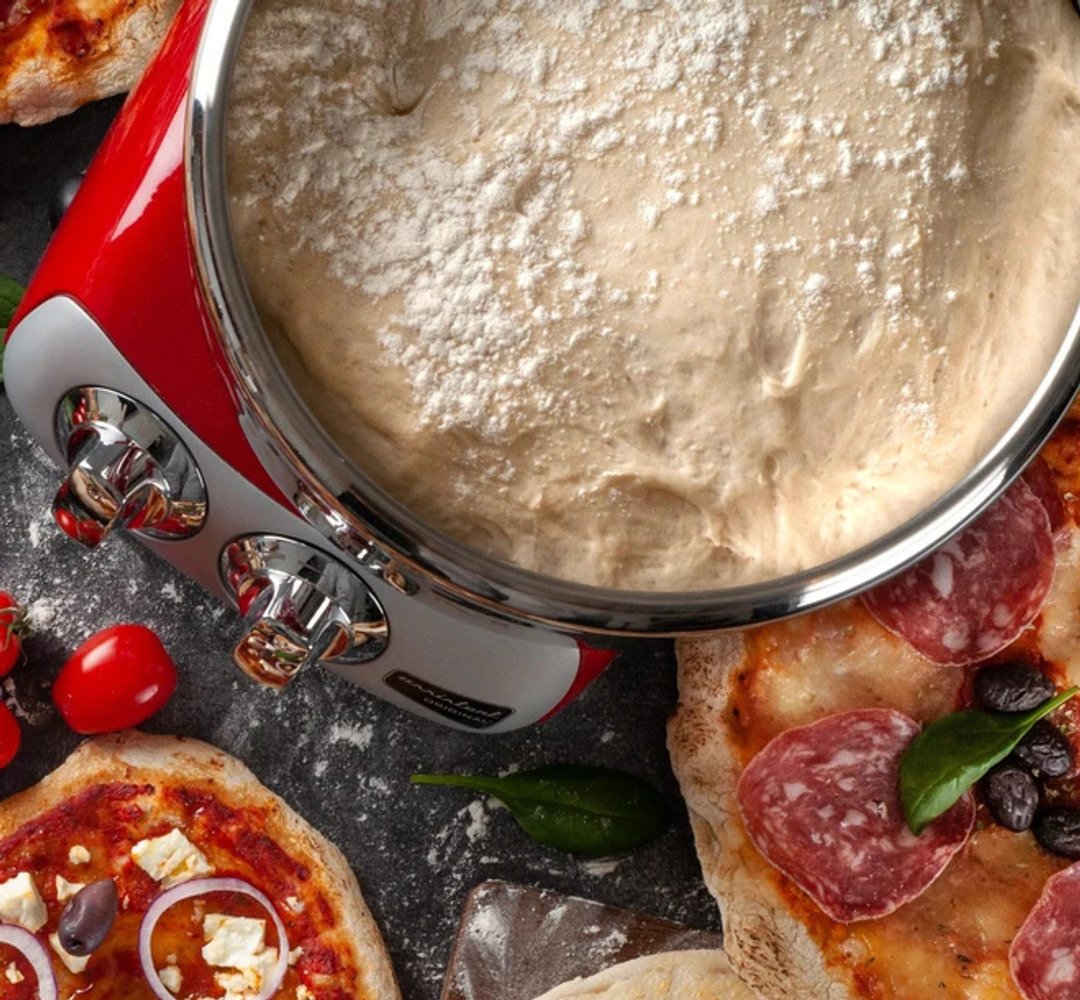 Prepared pizza dough in the Ankarsrum Assistent on a kitchen countertop, with freshly baked pizzas on the side.