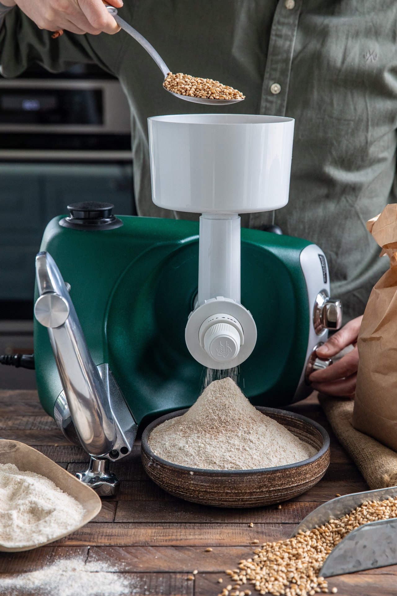 A person scooping grain into the grain mill attached to an Ankarsrum stand mixer.