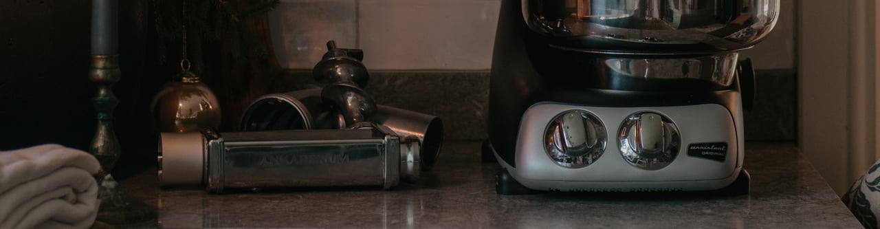 Ankarsrum Assistent Original keittiötasolla valoisassa keittiössä. Koneen vieressä on pastalisäosa, joka korostaa laitteen monipuolisuutta kotitekoisen pastan valmistuksessa.