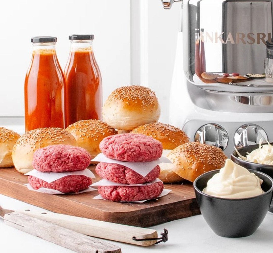 A wooden cutting board topped with hamburgers next to jars of ketchup and buns.