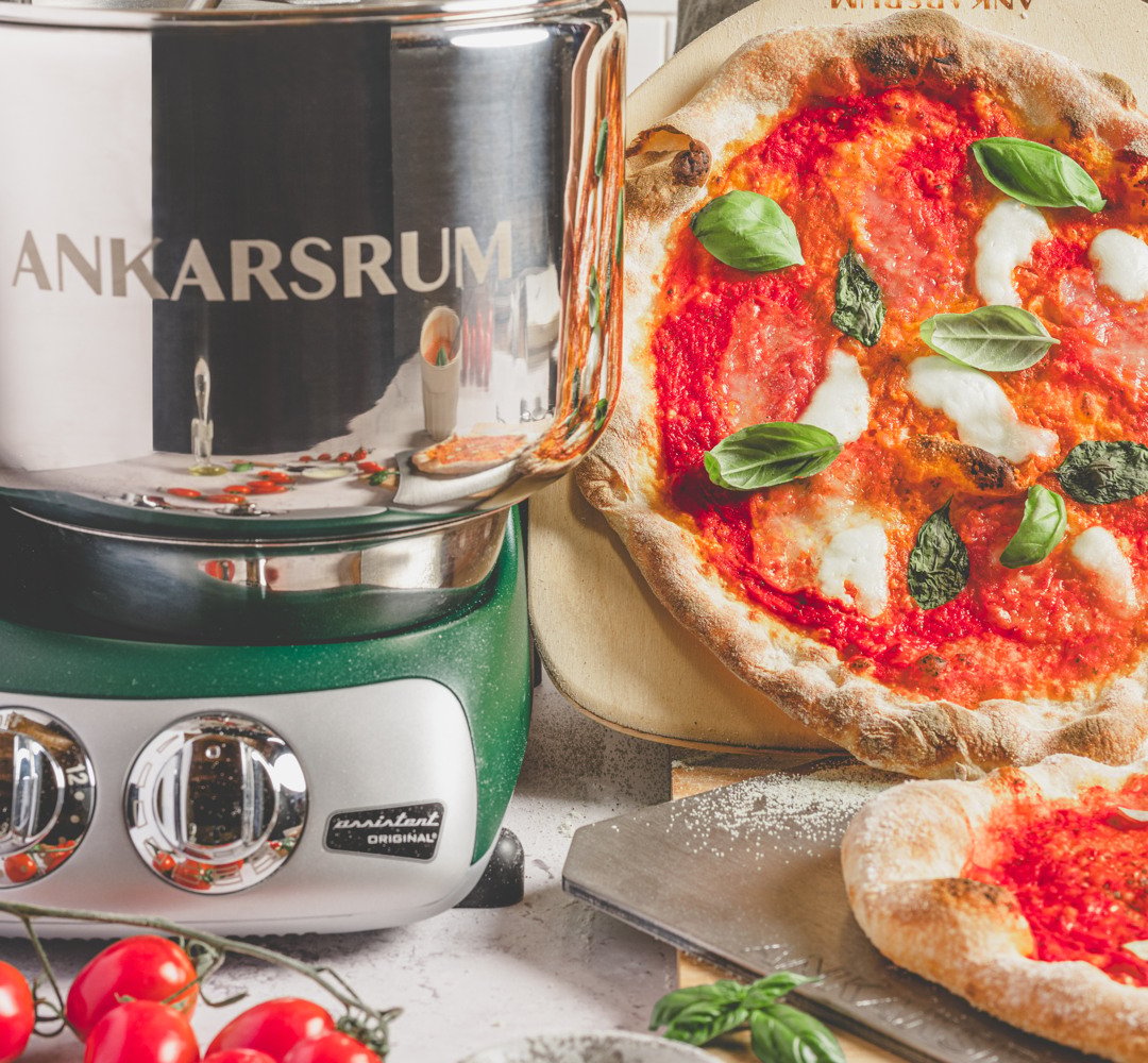 En pizza som ligger på en panna på en bänk. Ankarsrum assistent vid sidan.