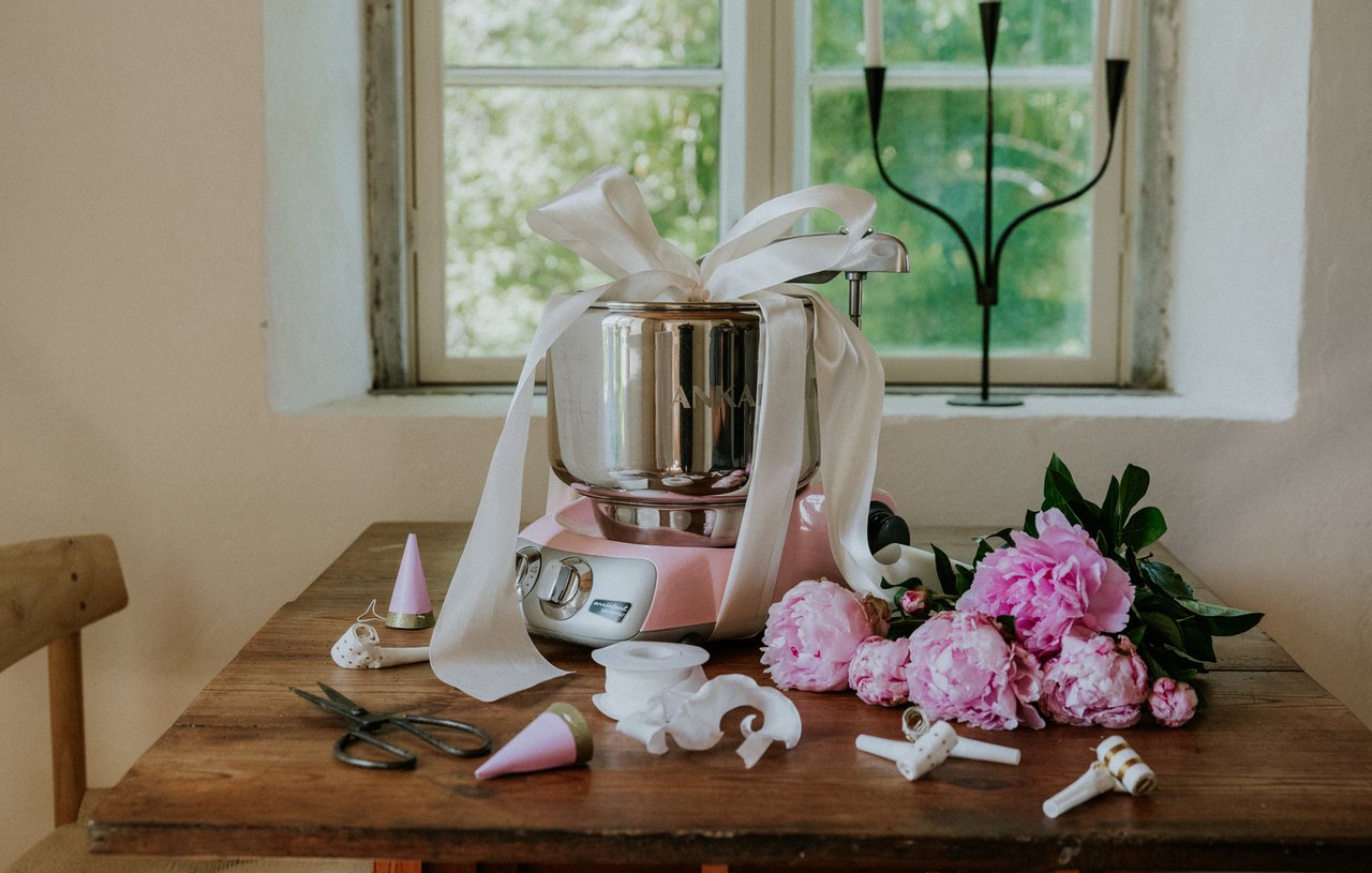 En kvinna dekorerar cupcakes med en spritspåse. På bordet står en Ankarsrum Assistent Original i Cloudy Pink, monterad med en vispskål i rostfritt stål.