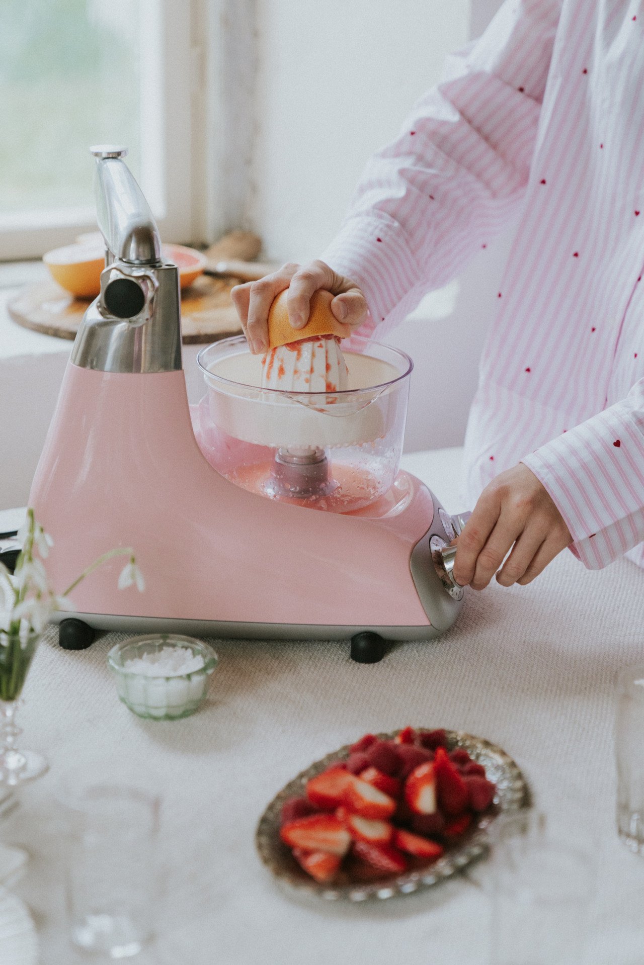 En Ankarsrum Assistent Original i molnig rosa, utrustad med Citruspress-tillbehöret, pressar färska apelsiner på ett köksbord bredvid en tallrik med jordgubbar.