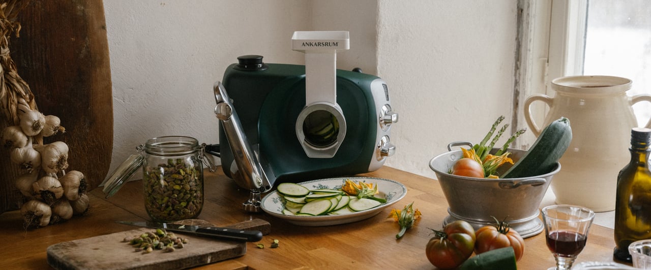 En Ankarsrum Assistent Original i Skogsgrön, utrustad med grönsakskniven, står på en träarbetsbänk i köket omgiven av färska grönsaker och skivad zucchini.