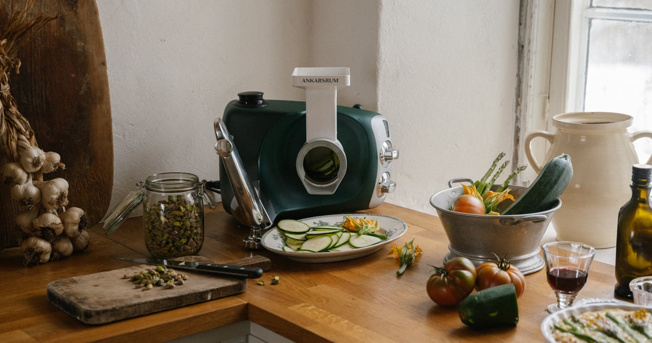 En Ankarsrum Assistent Original i Skogsgrön, utrustad med grönsakskniven, står på en träarbetsbänk i köket omgiven av färska grönsaker och skivad zucchini.