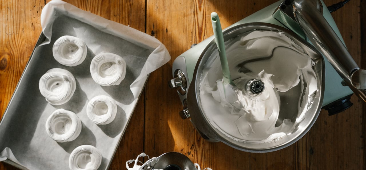 Ett träbord med en Ankarsrum-mixer fylld med marängsmet, bredvid en bakplåt med spritsade maränger.