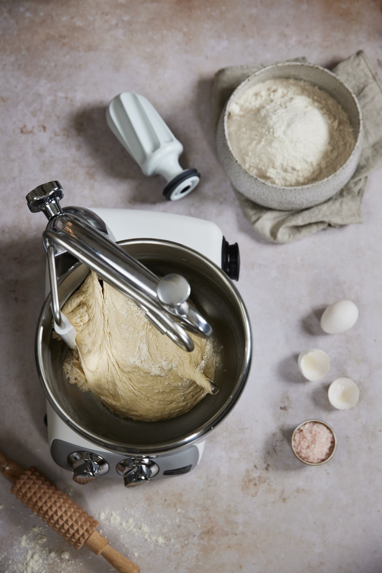 A mixer with a bowl of dough. Eggs, flour and a dough roller beside.