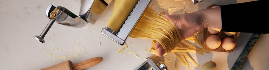 Fresh homemade pasta being prepared with Ankarsrums pasta rollers, surrounded by pasta dough, eggs, and flour on a kitchen worktop.