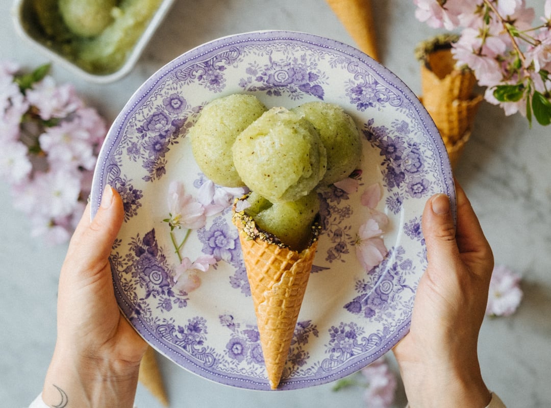 En person som håller en tallrik med en kiwi-sorbetkon. I bakgrunden syns rosa blommor, tillsammans med ett bord som har en skål med kvarvarande glass.