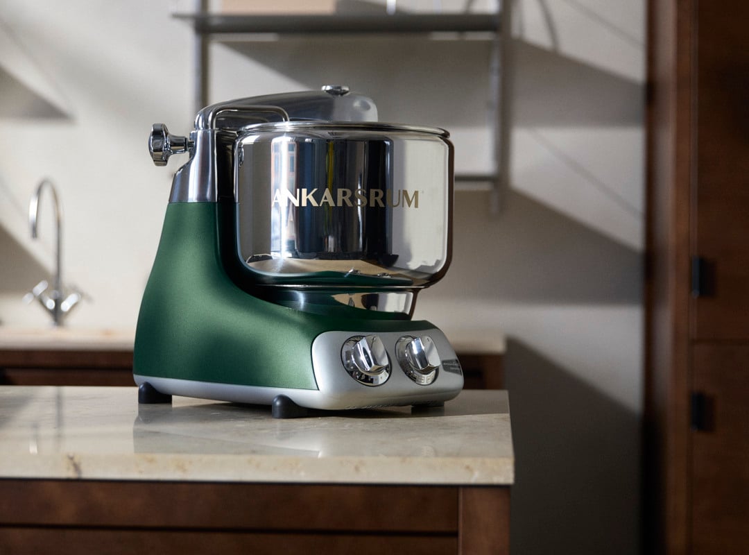 An Ankarsrum Assistent Original in color Forest Green with a stainless steel bowl on a kitchen counter, illuminated by warm sunlight.