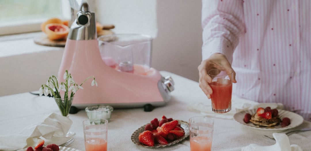 Ett dukat frukostbord med små pannkakor dekorerade med hallon och jordgubbar, glas med rosafärgad juice samt en tallrik med färska bär. I bakgrunden står en Ankarsrum Assistent Original i färgen Cloudy Pink, och en person klädd i en rosa skjorta håller ett glas juice. På bordet finns också en liten vas med vita blommor, samt bestick och vita tygservetter som skapar en ljus och inbjudande atmosfär.