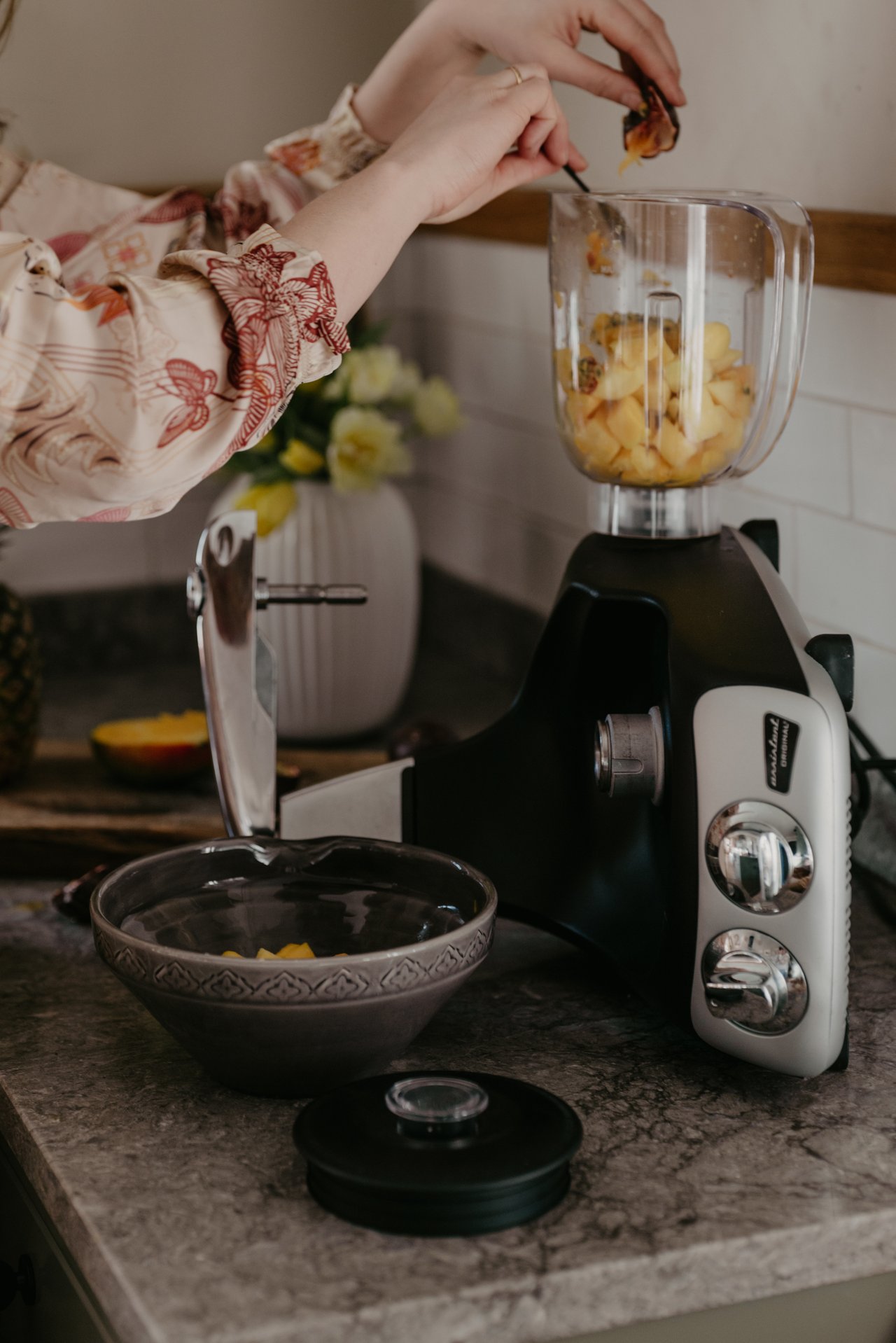 A woman uses the Blender attachment on an Ankarsrum Assistent Original in Harmony Beige to prepare a fruit smoothie in a bright kitchen.
