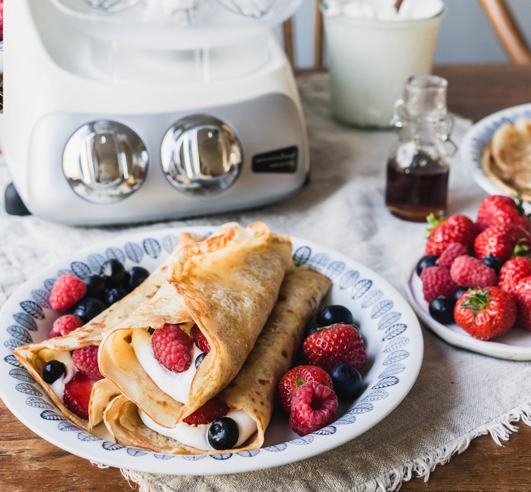 A plate of freshly cooked crêpes topped with berries and whipped cream, with an Ankarsrum Assistent in the background.