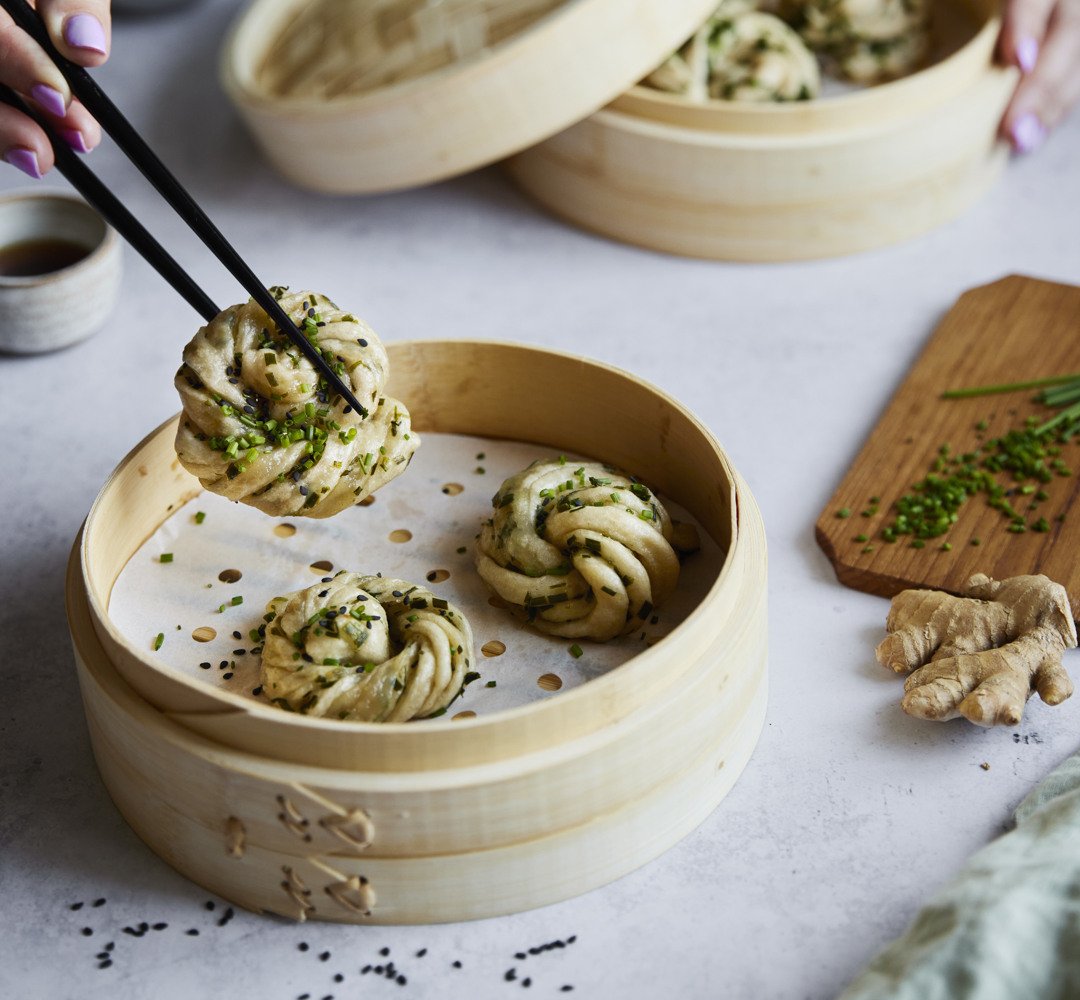 Bambukori pöydällä täynnä mantou-sämpylöitä. Henkilö poimii mantoun syömäpuikoilla.