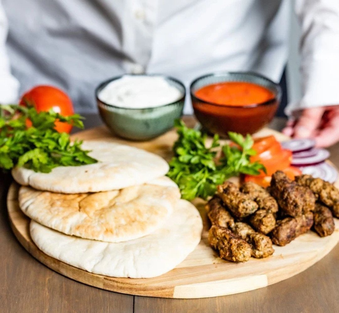 A large cutting board with cevapcici, pita bread, and various side dishes.