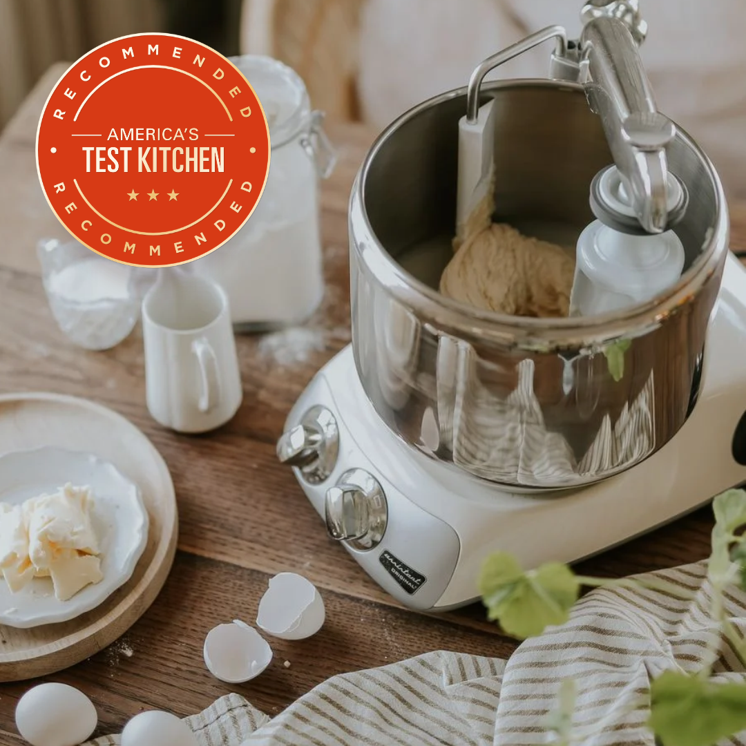 An Ankarsrum Assistent Original on a wooden table, surrounded by ingredients.