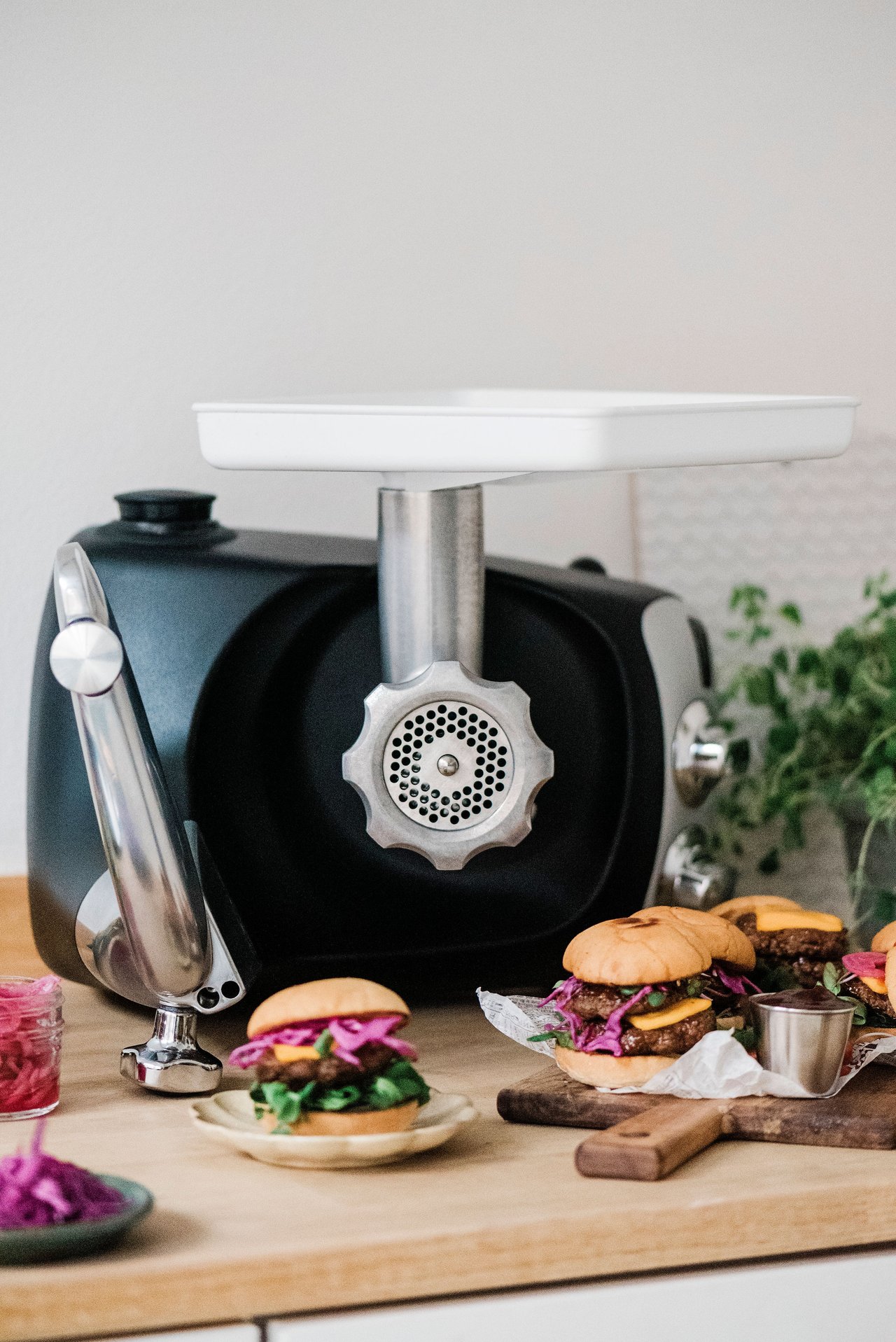 En skärbräda med färdiga hamburgare och bröd. En Ankarsrum Assistent och en köttkvarn i bakgrunden.