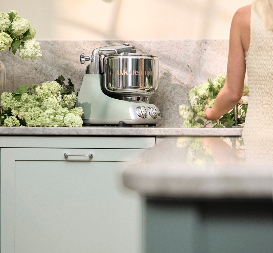 An Ankarsrum Assistent Original in Nordic Sage stands on a kitchen worktop surrounded by green hydrangeas, while a woman arranges flowers nearby.