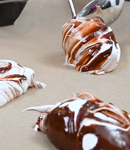 En kvinna som skedar smält choklad tillsammans med maräng på en bakplåt.
