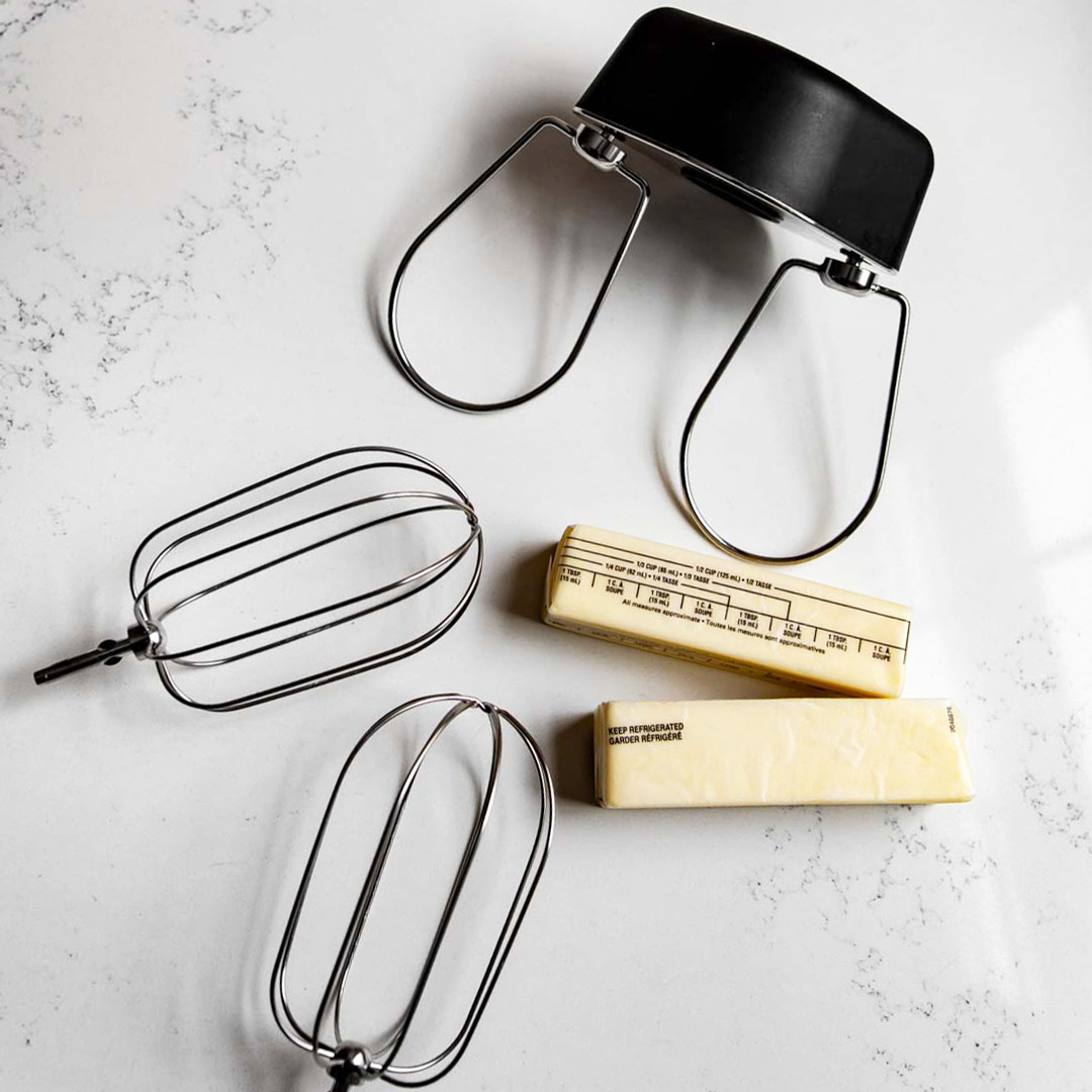 Ankarsrum Premium Beater Set vist på en lys marmorbordplade med store ballonpiskeris til hurtigere piskning og lettere, luftigere teksturer, robuste kagepiskere til blanding af tærte-, wienerbrøds- og småkagedeje op til 1 kg (2 lb), et sort tilbehørshus samt to pakker smør. Premium-tilbehør til køkkenmaskinen, designet til at sikre ensartet blanding og bagerikvalitet i dine bageresultater.