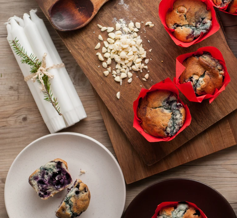 Freshly baked blueberry white chocolate chip muffins in red tulip liners on a wooden cutting board, made with Ankarsrum stand mixer.