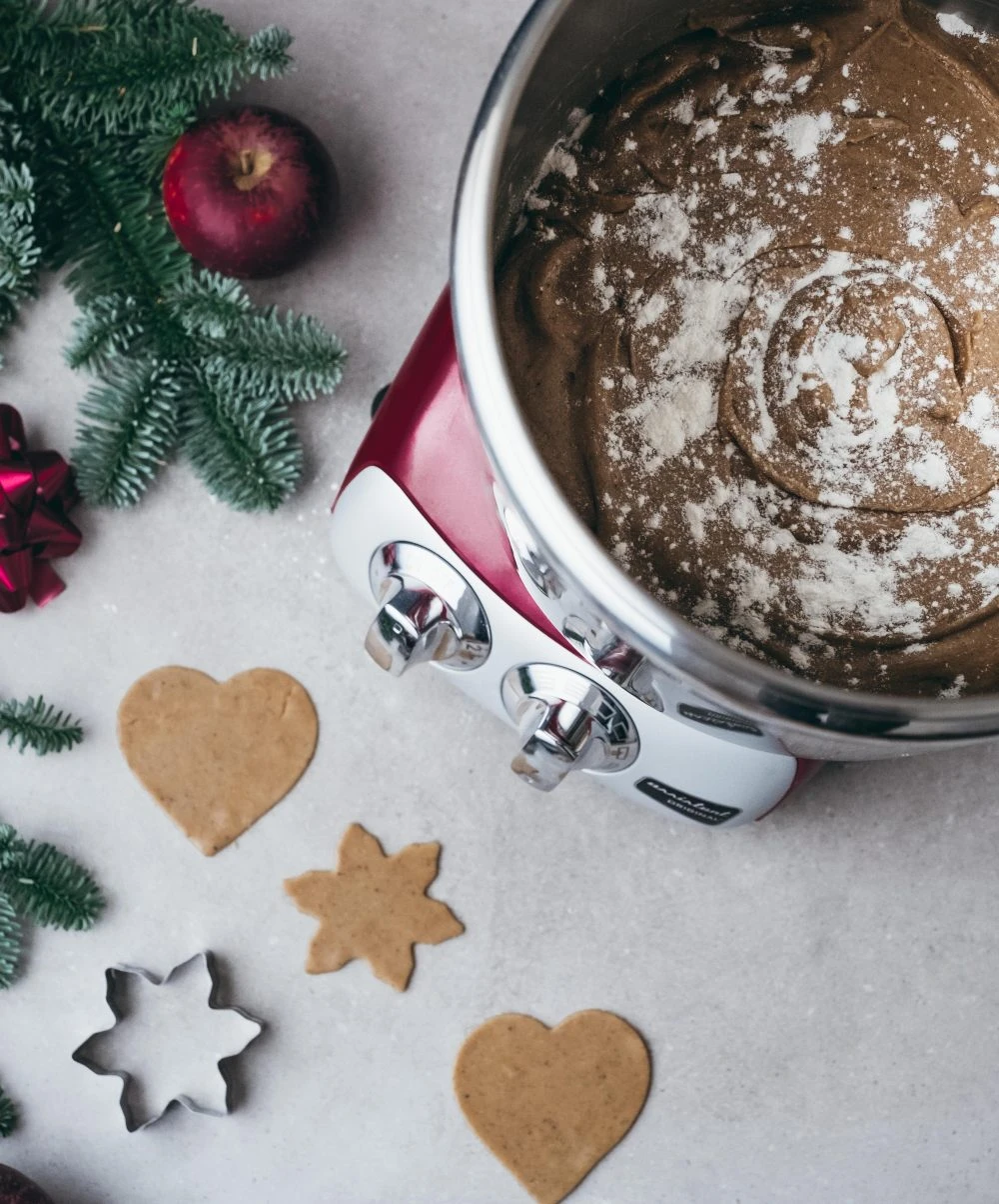 Ankarsrum red stand mixer with gingerbread dough, heart and star-shaped cookie cutouts, pine branches, and Christmas ornaments.