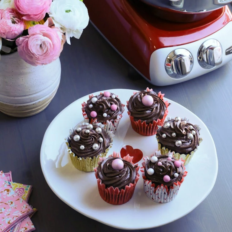 Seven chocolate cupcakes with pearl decorations on a white plate, beside an Ankarsrum red stand mixer and pink flowers.